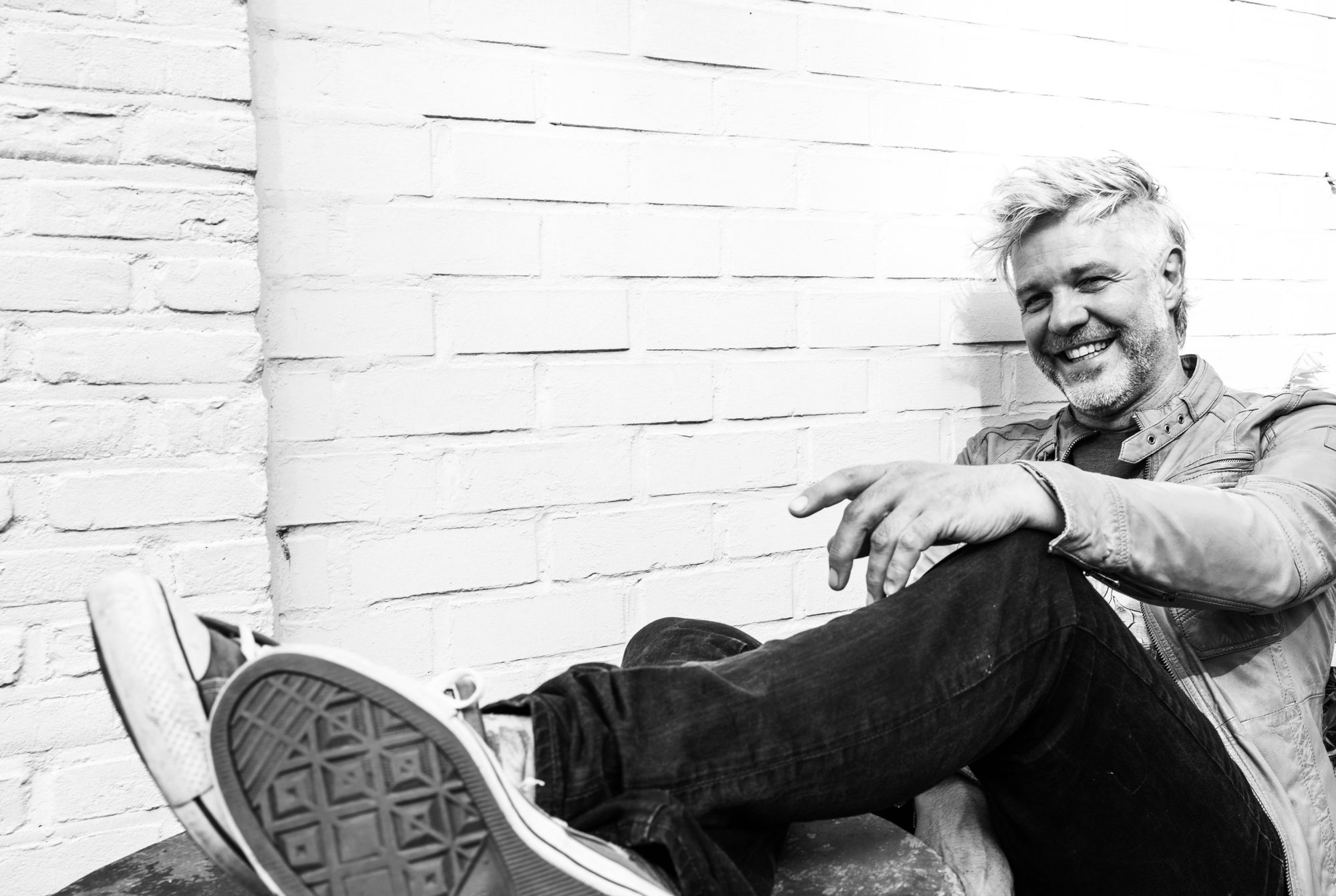 Black and white photo of Robin Dhondt laughing, sitting against a white brick wall with his worn sneakers up in front of the camera