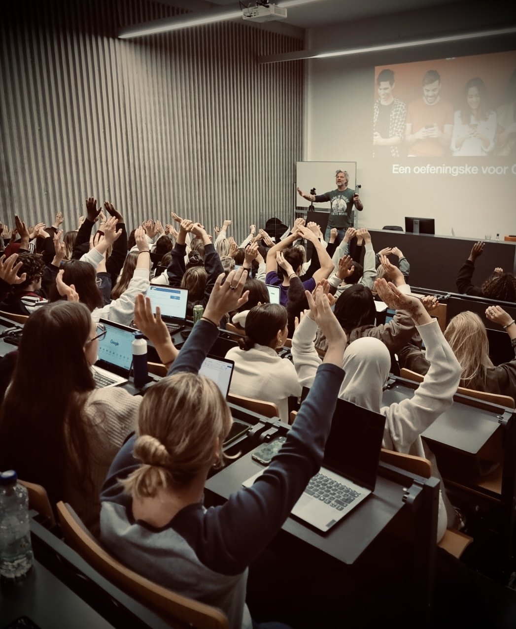 A full lecture hall with everyone raising their hands during an exercise, Robin small at the front next to the screen